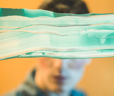 visuell unterstützende Graphik zu LV Trans*, Trans*, Trans*phobie  Bildinhalt: Unscharfe Person vor gelbem Hintergrund, die Augen durch breite, fließende Farbbänder in Türkis und Weiß horizontal verdeckt; wirkt wie eine visuelle Barriere und Symbol für Unsichtbarmachung.